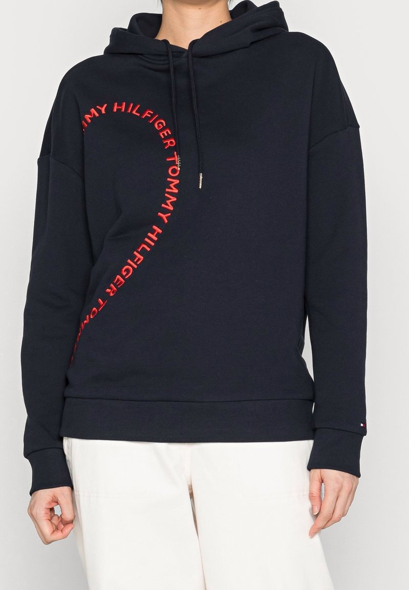 Personne portant un sweat à capuche noir avec un texte rouge incurvé "Tommy Hilfiger" sur le devant et un pantalon blanc, visible du cou jusqu'aux hanches.