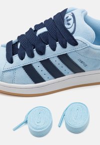 Sapatilhas de camurça azul claro com design em três riscas azul marinho, atacadores planos azul marinho e sola branca. Inclui dois atacadores de reposição azul claro.