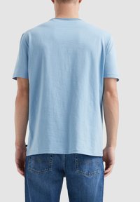Jasnoniebieska bawełniana koszulka z okrągłym dekoltem i krótkimi rękawami, noszona na odwrót, zestawiona z niebieskimi dżinsami z tylnymi kieszeniami.