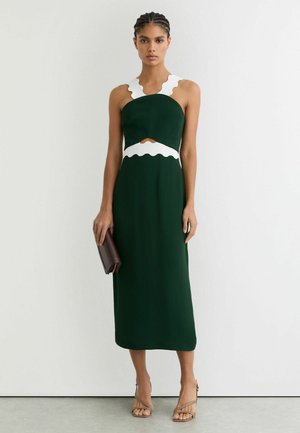 Femme en robe midi sans manches vert foncé avec bordure blanche ondulée, tenant une pochette marron foncé et portant des sandales beiges.
