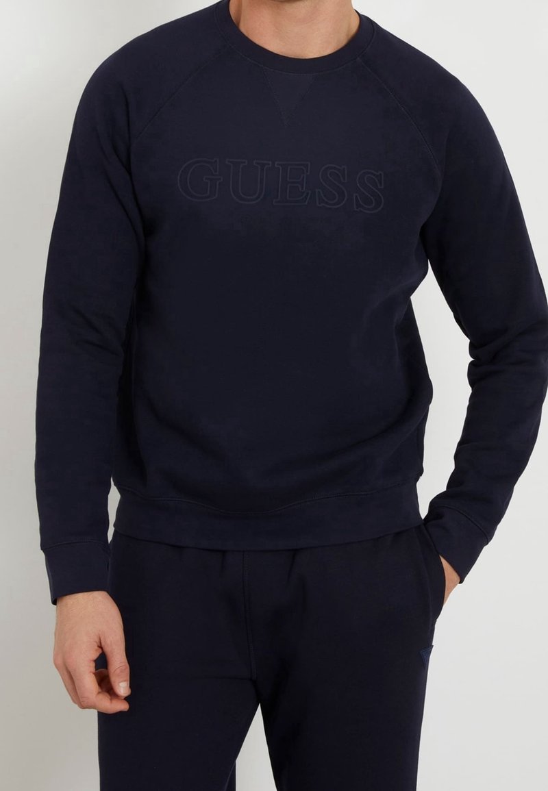 Man som bär en svart sweatshirt och matchande svarta byxor, med en diskret "GUESS"-logotyp på bröstet, stående med en hand i fickan.