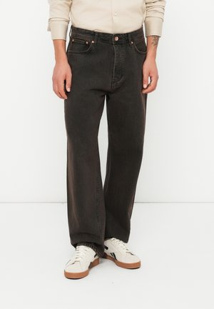 Homme portant un jean noir à jambes larges, une chemise beige à manches longues et des baskets blanches, debout devant un fond blanc uni.