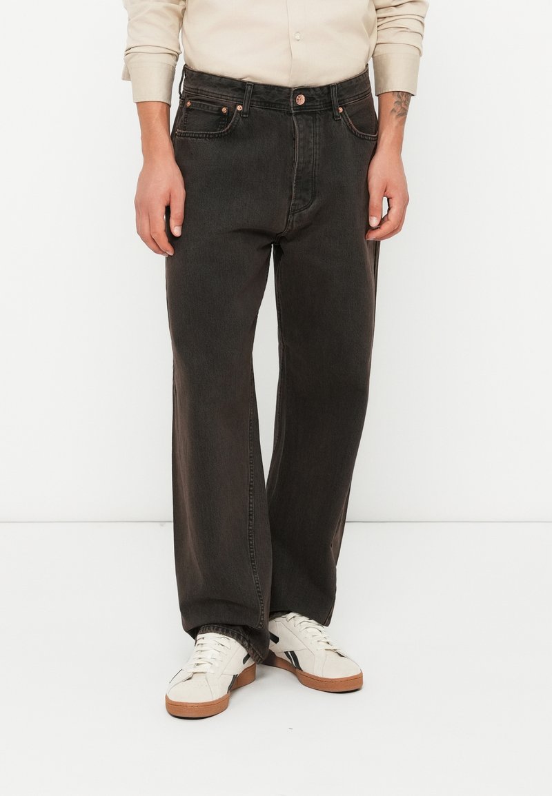 Homme portant un jean noir à jambes larges, une chemise beige à manches longues et des baskets blanches, debout devant un fond blanc uni.
