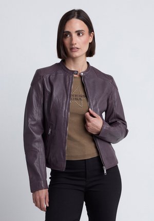 Vrouw met kort donker haar die een paarse leren jas draagt over een bruin shirt met sterontwerp en een zwarte broek, staand tegen een grijze achtergrond.