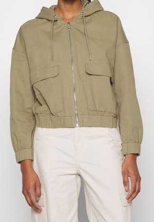 Giacca con cappuccio verde oliva con chiusura a zip frontale, vita elasticizzata e due tasche sul petto. Tessuto strutturato con polsini arricciati.