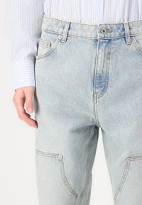 Jeans de denim azul con un acabado desgastado, que cuentan con múltiples bolsillos, costuras visibles y un cierre de botón en la cintura.