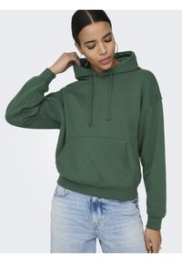Femme en sweat à capuche vert ajustant sa capuche avec la main, portant un jean bleu clair et des boucles d'oreilles anneau, sur un fond clair uni.