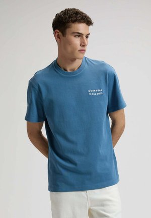 Junger Mann mit lockigem Haar, der ein blaues T-Shirt mit dem Schriftzug "WORKWEAR IS OUR SOUL" trägt und mit den Händen hinter dem Rücken vor einfarbigem Hintergrund steht.