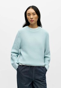 Jersey de punto azul claro con cuello redondo acanalado y mangas largas, llevado sobre una camiseta blanca, combinado con pantalones vaqueros oscuros.