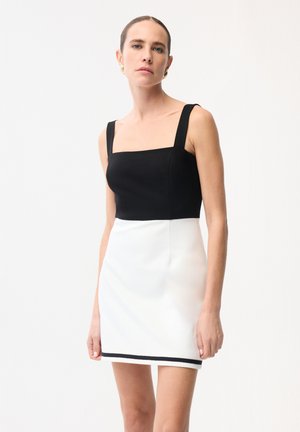 Donna con indosso un top nero senza maniche e una gonna bianca con un bordo nero all'orlo, in piedi davanti a uno sfondo chiaro e uniforme.