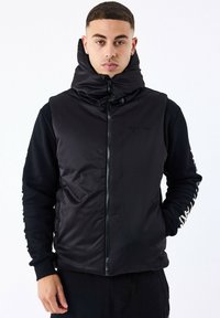 Sort polstret vest med høj krave, lynlåslukning og sidelommer; har sorte ærmer med hvid tekst langs armen. Enkel og funktionel design.