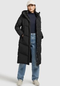 Donna che indossa un lungo piumino nero, jeans azzurri, sneaker bianche, dolcevita blu navy e un cappellino chiaro e texture, in piedi con le mani in tasca.