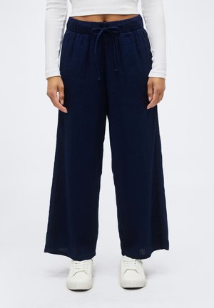 Kvinde iført marineblå bukser med vide ben, elastisk talje og snøre, hvid cropped top med lange ærmer og hvide sneakers.