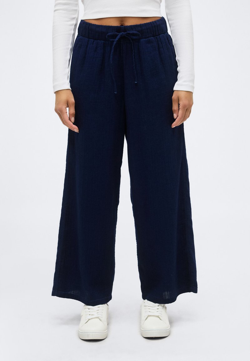 Vrouw draagt marineblauwe wijde broek met elastische tailleband en trekkoord, wit cropped top met lange mouwen en witte sneakers.