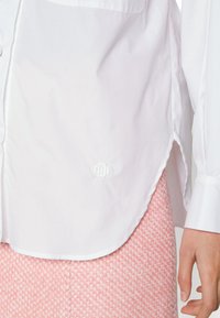 Camicia bianca con bottoni realizzata in tessuto liscio, caratterizzata da un sottile dettaglio ricamato sull'orlo e un orlo curvo, abbinata a una gonna rosa con texture.