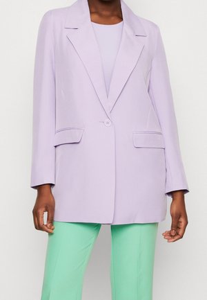 Femme portant un blazer violet clair à un bouton avec poches à rabat par-dessus un top assorti et un pantalon vert menthe sur un fond uni.