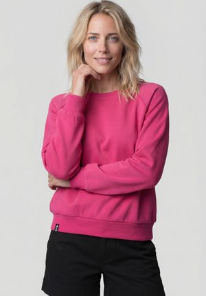 Blonde Frau lächelt, trägt einen knallpinken Pullover und schwarze Shorts, posiert mit einer Hand nahe am Kinn vor hellem Hintergrund.