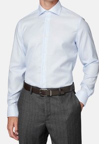 Uomo che indossa una camicia a righe leggera blu chiaro infilata in pantaloni grigio scuro a righe con una cintura marrone scuro, con la mano in tasca.