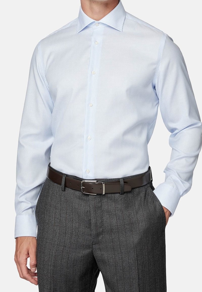 Uomo che indossa una camicia a righe leggera blu chiaro infilata in pantaloni grigio scuro a righe con una cintura marrone scuro, con la mano in tasca.
