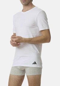 T-shirt bianco a maniche corte in cotone con scollo rotondo e logo Adidas nero sul lato sinistro, abbinato a pantaloni boxer grigi.
