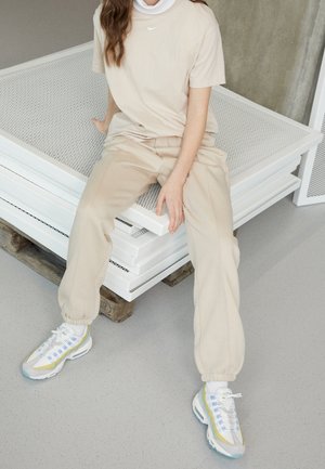 Personne assise sur des panneaux métalliques blancs empilés, vêtue d'une tenue Nike beige et de baskets blanches, bleues et jaunes sur un sol gris.