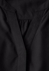 Blouse noire avec un décolleté en V, des détails froncés, une texture lisse et des coutures contrastantes le long des bords.