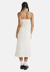 Vestido maxi blanco sin mangas con estampado floral y espalda fruncida, que presenta tirantes finos y un ajuste relajado, combinado con zapatos negros y calcetines blancos.