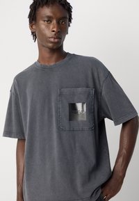 En ung mann med korte dreads iført en falmet mørkegrå oversized T-skjorte med en lommepåse, som har et blankt svart kvadrat og tekst.