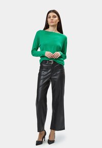 Grünes Langarm-Oberteil mit weitem Ausschnitt, kombiniert mit hochgeschnittenen schwarzen Lederimitat-Hosen. Schwarze hohe Absätze runden das Outfit ab.