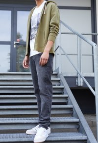 Sudadera de cierre en verde oliva, camiseta gráfica blanca, jeans gris oscuro y zapatillas blancas. Textura de tela suave y corte casual, de pie en escalones de metal.