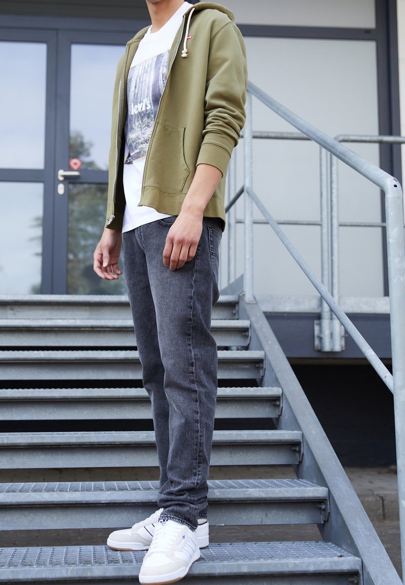 Sudadera de cierre en verde oliva, camiseta gráfica blanca, jeans gris oscuro y zapatillas blancas. Textura de tela suave y corte casual, de pie en escalones de metal.