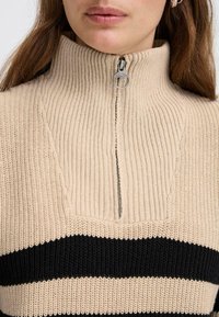 Beige und schwarz gestreifter Pullover mit hohem geripptem Kragen und halber Reißverschluss. Texturiertes Strickgewebe. Akzent aus silbernem Reißverschluss-Zubehör.