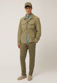 Veste en daim vert olive clair avec deux poches avant, fermeture à boutons et couture ton sur ton, associée à une chemise à motifs et un pantalon olive.