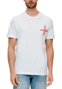 Camiseta de algodón blanca con un patrón geométrico texturizado rojo y blanco en el área del lado izquierdo del pecho. Diseño de mangas cortas y cuello redondo.