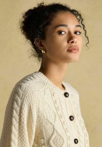 Krémový pletený kardigán s vrúbkovou textúrou, s tmavými gombíkmi a okrúhlym výstrihom. Jednoduchý dizajn, mäkká tkanina, teplý vzhľad.