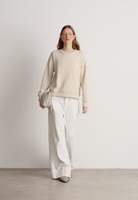 Beżowy sweter z fakturowanym przodem, zestawiony z białymi szerokimi spodniami. Modelka ma w dłoni jasną torebkę kopertową i nosi szare buty.