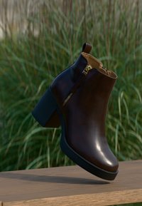 Dark brown leather ankle boot with side zipper and block heel, displayed on wooden surface against blurred tall green grass background.