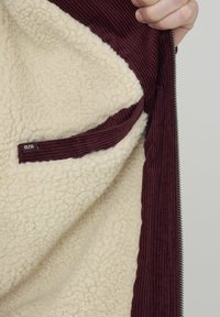 Veste à fermeture éclair extérieure bordeaux avec une doublure douce en sherpa crème et des accents en velours côtelé texturé. Comprend une étiquette BZB sur la poche.