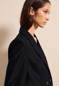 Blazer negro a medida con solapa tipo muesca, botones dobles y hombros estructurados. La tela tiene un brillo sutil y una textura suave.