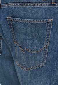 Τζιν παντελόνι με σκούρο μπλε ξέβγαλμα, με τσέπη στην πίσω πλευρά με πορτοκαλί ραφές αντίθεσης και ένα λεπτό κυρτό διακοσμητικό στοιχείο.