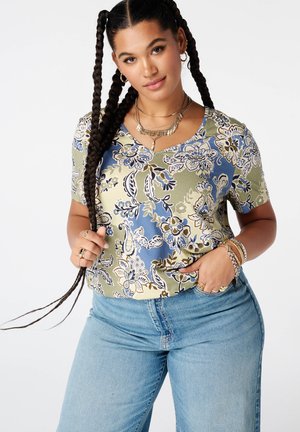 Vrouw met lang gevlochten haar draagt een korte mouwen blouse met bloemenprint, lichtblauwe spijkerbroek en gelaagde kettingen tegen een witte achtergrond.