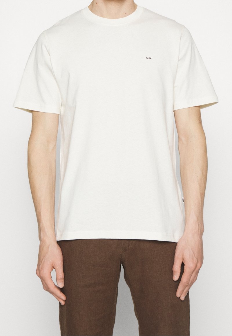 Krémové bavlněné tričko s krátkým rukávem, kulatým výstřihem a malým černým logem "w.w." na hrudi. V kombinaci s hnědými kalhotami.