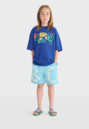 Bambino in piedi con le mani in tasca, indossa una maglietta blu con stampa floreale, pantaloncini azzurri con quadrati bianchi e sandali a ciabatta blu-verdi.