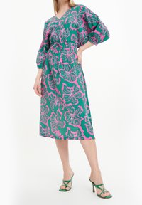 Mujer vistiendo un vestido midi floral en verde y rosa, con mangas abullonadas de 3/4 y sandalias de tacón alto verdes con tiras, posando con una mano en la cadera.