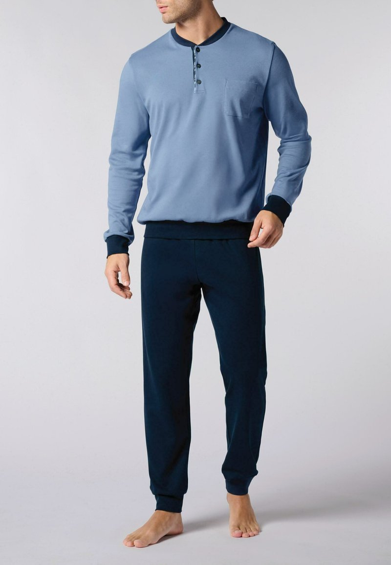 Set di abbigliamento da casa per uomo che presenta una maglia a maniche lunghe di colore azzurro chiaro con collo a bottoni e tasca sul petto, abbinata a pantaloni tapered di colore blu scuro.