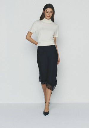 Woman wearing a white short-sleeve top and black midi skirt with lace hem, standing against a plain light gray background.