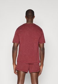 Camiseta de algodón burdeos con cuello redondo y mangas cortas, que presenta un pequeño logo en la parte posterior. Tejido suave y texturizado con un corte relajado.