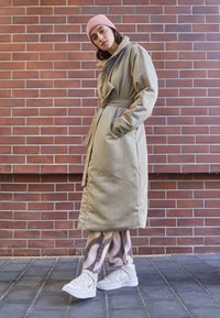 Beige pitkä takki, jossa on vyötärölle sidottu vyö, sivutaskut, yhdistetty kuvioituihin housuihin ja vaaleisiin lenkkareihin. Vaaleanpunainen neulottu pipo päässä.