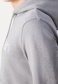 Personne portant un sweat à capuche gris clair avec un logo blanc partiel sur la poitrine, une couture d'épaule visible et une capuche reposant sur le dos.