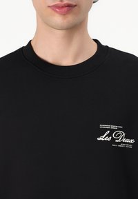 Czarny sweatshirt wykonany z miękkiego materiału, z ribbowanym dekoltem, małym białym logo i napisem po lewej stronie z przodu.